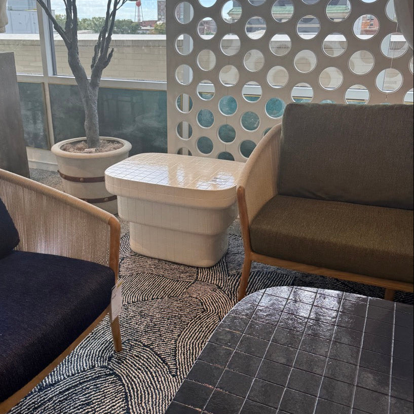 A modern outdoor lounge seating area featuring two wooden armchairs with dark blue and olive green cushions, a small white side table, a black tiled coffee table, and a decorative circular screen wall.