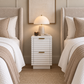 A modern white ribbed nightstand with gold handles sits between two matching beds in a neutral bedroom setting. A lit white dome lamp and a small candle rest on top of the nightstand, which is centered under a framed wall art piece.