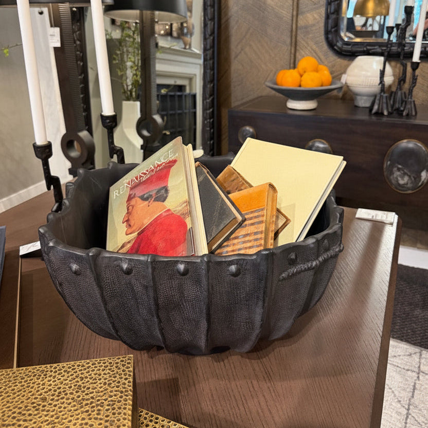 Decorative metal bowl with books and a tag on a wooden surface, with mirrors and lamps in the background.