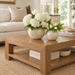 A modern square wooden coffee table in a bright living room, styled with two textured cream ceramic vases containing white hydrangeas and tulips.