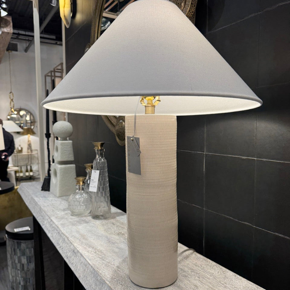 Table lamp with a gray shade on a display stand against a black tiled wall.