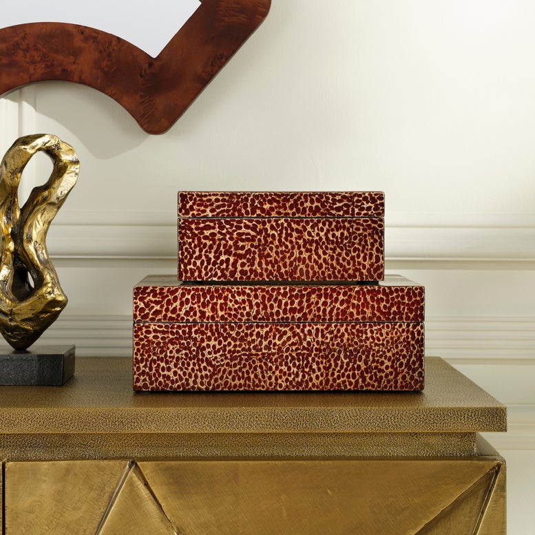Two stacked rectangular decorative boxes with a red and beige animal-print pattern, displayed on a metallic gold console table against an off-white wall.