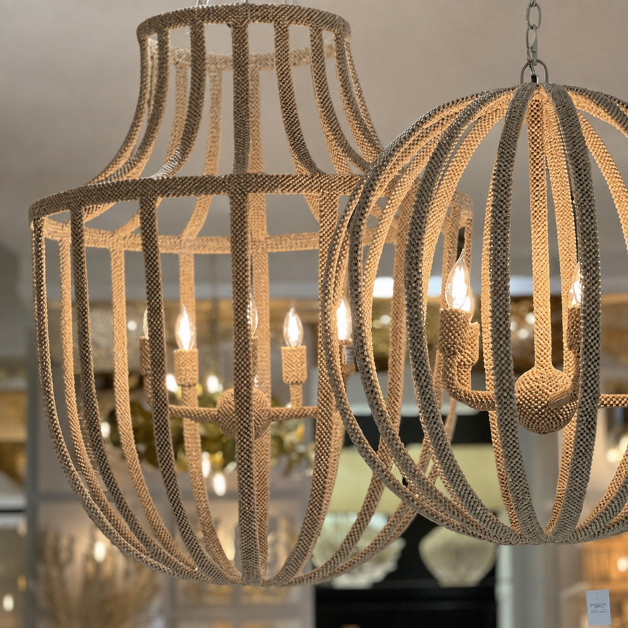 Two woven rope chandeliers hanging in a softly blurred showroom. One is a tall, tiered cage-style chandelier with candle-style lights, and the other is a spherical, open-frame chandelier also lit with multiple candle bulbs.