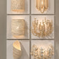Six decorative wall sconces displayed on a white grid wall, each mounted on a square panel. The fixtures are in warm metallic gold and cream tones, including a scalloped shell-like shade, a beaded cylindrical wall light, a floral chandelier-style sconce, a sculptural shell form, and a leafy wreath-style light.