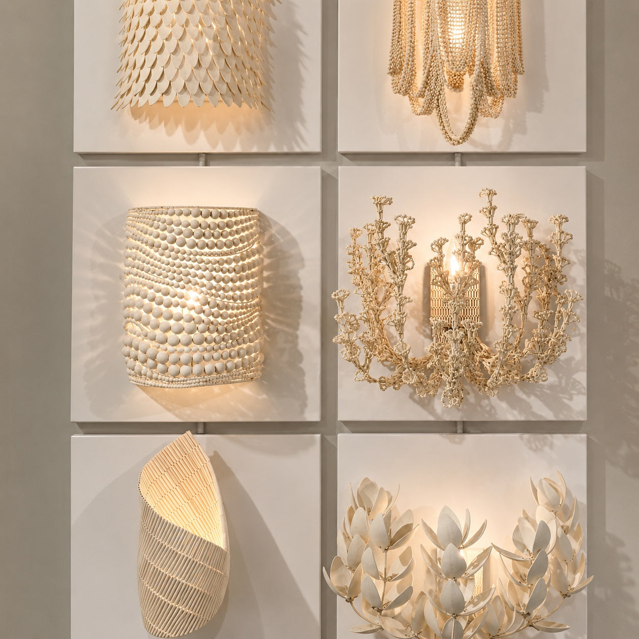 Six decorative wall sconces displayed on a white grid wall, each mounted on a square panel. The fixtures are in warm metallic gold and cream tones, including a scalloped shell-like shade, a beaded cylindrical wall light, a floral chandelier-style sconce, a sculptural shell form, and a leafy wreath-style light.