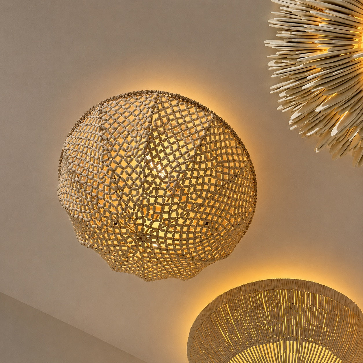 A spherical woven pendant chandelier with a geometric openwork pattern, glowing warm yellow light against a neutral beige ceiling. Part of a larger spiky chandelier and another woven lamp are visible at the edges.