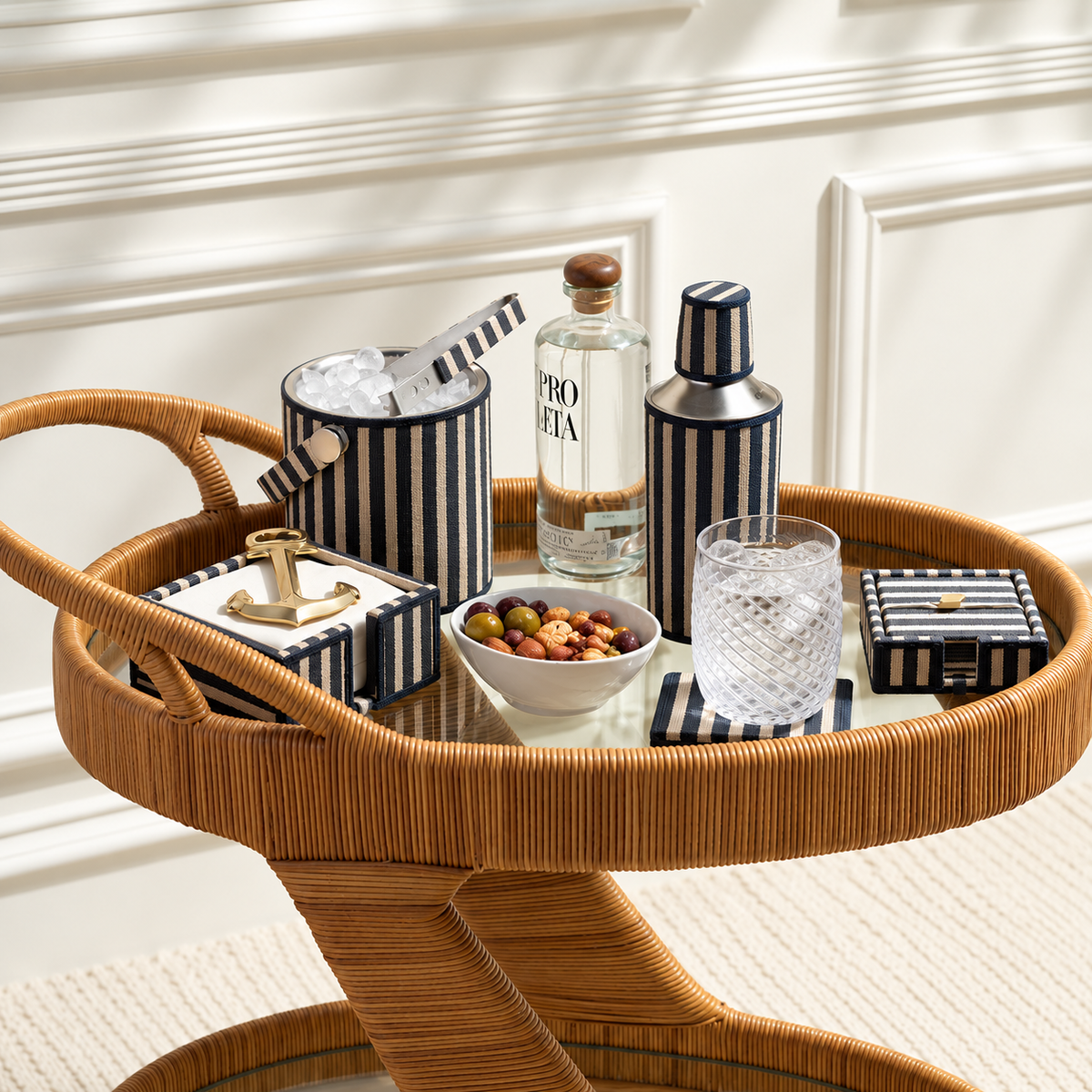 A round woven rattan bar cart styled with cocktail accessories, including a striped bucket with a straw, a shaker, a bottle of Prosecco, a glass, and matching striped boxes, set in front of a white paneled wall.
