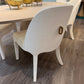 A modern white dining chair with a curved backrest and a gold keyhole accent, shown in a dining room setting on a gray wood floor next to a light wood table.