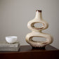 A modern, abstract ceramic vase with two stacked oval loops and a narrow neck, finished in a glossy cream glaze with brown speckled and mottled detailing. It is displayed on a dark wooden table next to a small white bowl on a stack of books, set against a neutral gray wall.