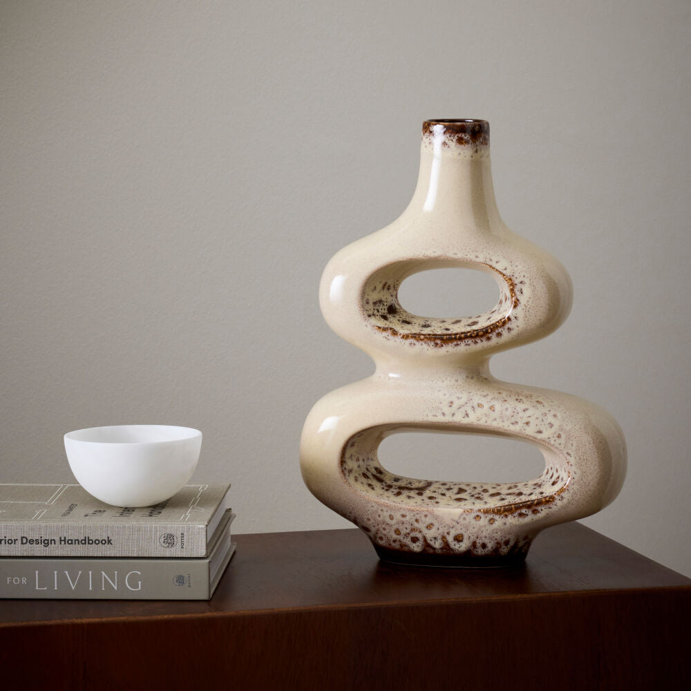 A modern, abstract ceramic vase with two stacked oval loops and a narrow neck, finished in a glossy cream glaze with brown speckled and mottled detailing. It is displayed on a dark wooden table next to a small white bowl on a stack of books, set against a neutral gray wall.