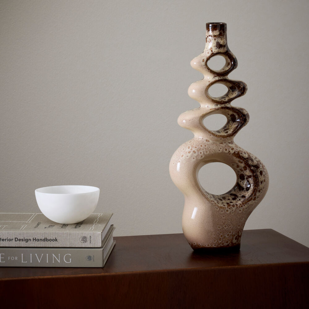 An abstract, sculptural ceramic vase with a glossy beige and dark brown speckled glaze stands on a dark wooden table next to a small white bowl stacked on two books against a neutral beige wall.