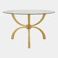 A round dining table featuring a clear glass top and a unique gold-colored metal base composed of five curved, ribbed legs that converge at the center, shown against a plain light gray background.