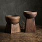Two dark brown wooden pedestal planters with a rounded bowl top and geometric base, displayed on a rustic wooden table against a dark gray textured background.