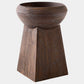A dark brown wooden pedestal planter with a wide, rounded bowl on top of a tapered geometric base, shown against a plain light gray background.