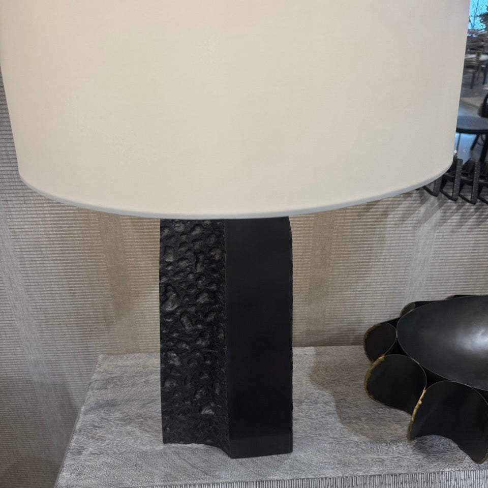 A modern table lamp with a large cream-colored drum shade and a black textured rectangular base, shown on a gray patterned side table.