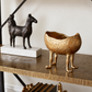 A textured metallic gold planter shaped like a three-legged animal stands on a dark wood console table next to a bronze horse sculpture on a white marble base.