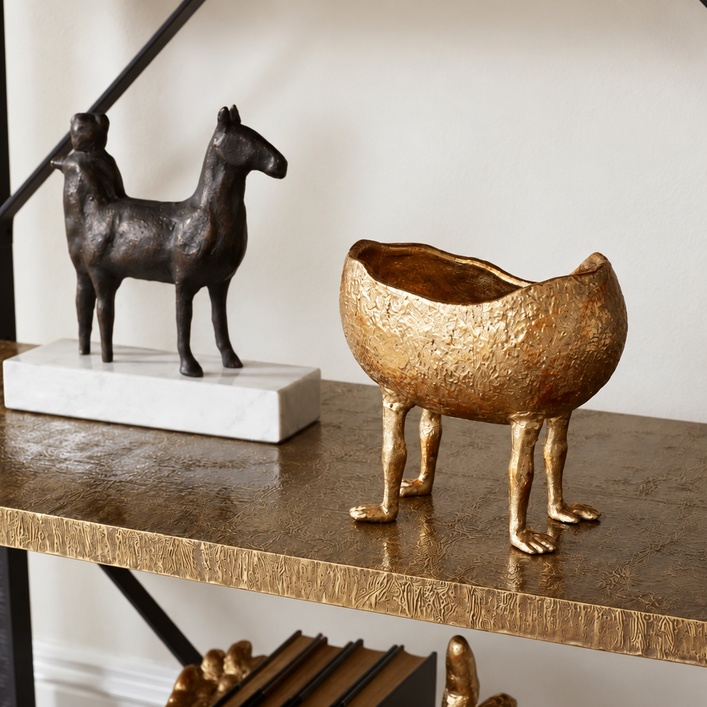 A textured metallic gold planter shaped like a three-legged animal stands on a dark wood console table next to a bronze horse sculpture on a white marble base.