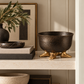 Textured black decorative bowl with sculptural gold feet styled on a neutral stone shelf alongside stacked books, a black marble tray, abstract artwork, and an olive branch arrangement in a modern luxury interior.