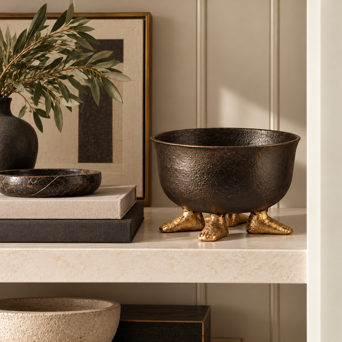 Textured black decorative bowl with sculptural gold feet styled on a neutral stone shelf alongside stacked books, a black marble tray, abstract artwork, and an olive branch arrangement in a modern luxury interior.