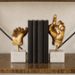 A pair of gold hand sculptures on white marble bases holding a set of three black books on a wooden shelf against a textured beige wall.