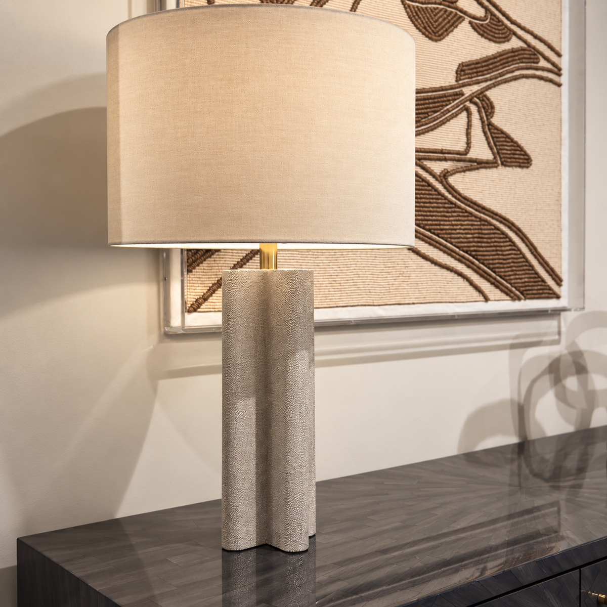 A modern table lamp with a textured beige fabric drum shade and a sculptural, fluted cylindrical base in a warm gray stone or concrete finish, shown on a dark wood console table in front of a framed abstract print.