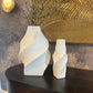 Two white abstract vases on a dark wooden surface with a textured wall in the background.