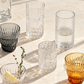 A set of textured glass tumblers and a small glass bowl arranged on a sunlit marble surface. The collection includes clear patterned tumblers, a dark gray ribbed tumbler, an amber/yellow tinted tumbler, and a small clear bowl with a lemon wedge and herb sprig.