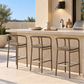 Three outdoor bar stools with woven seats and a weathered bronze finish, positioned at a stone kitchen island on a patio.