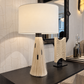A table lamp with a large white drum shade and a sculptural base made of cream-colored fibers, displayed on a dark wooden console table in a modern interior setting.