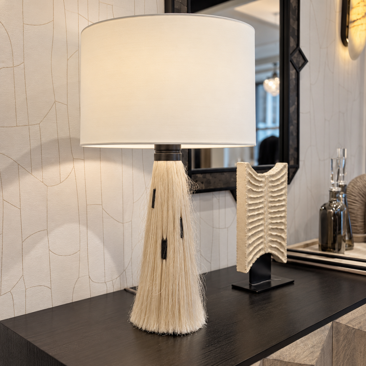 A table lamp with a large white drum shade and a sculptural base made of cream-colored fibers, displayed on a dark wooden console table in a modern interior setting.