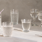 A collection of clear textured glass drinking vessels, including tumblers, a goblet, and a partially visible tumbler with a straw, arranged on a light stone surface against a neutral gray background.