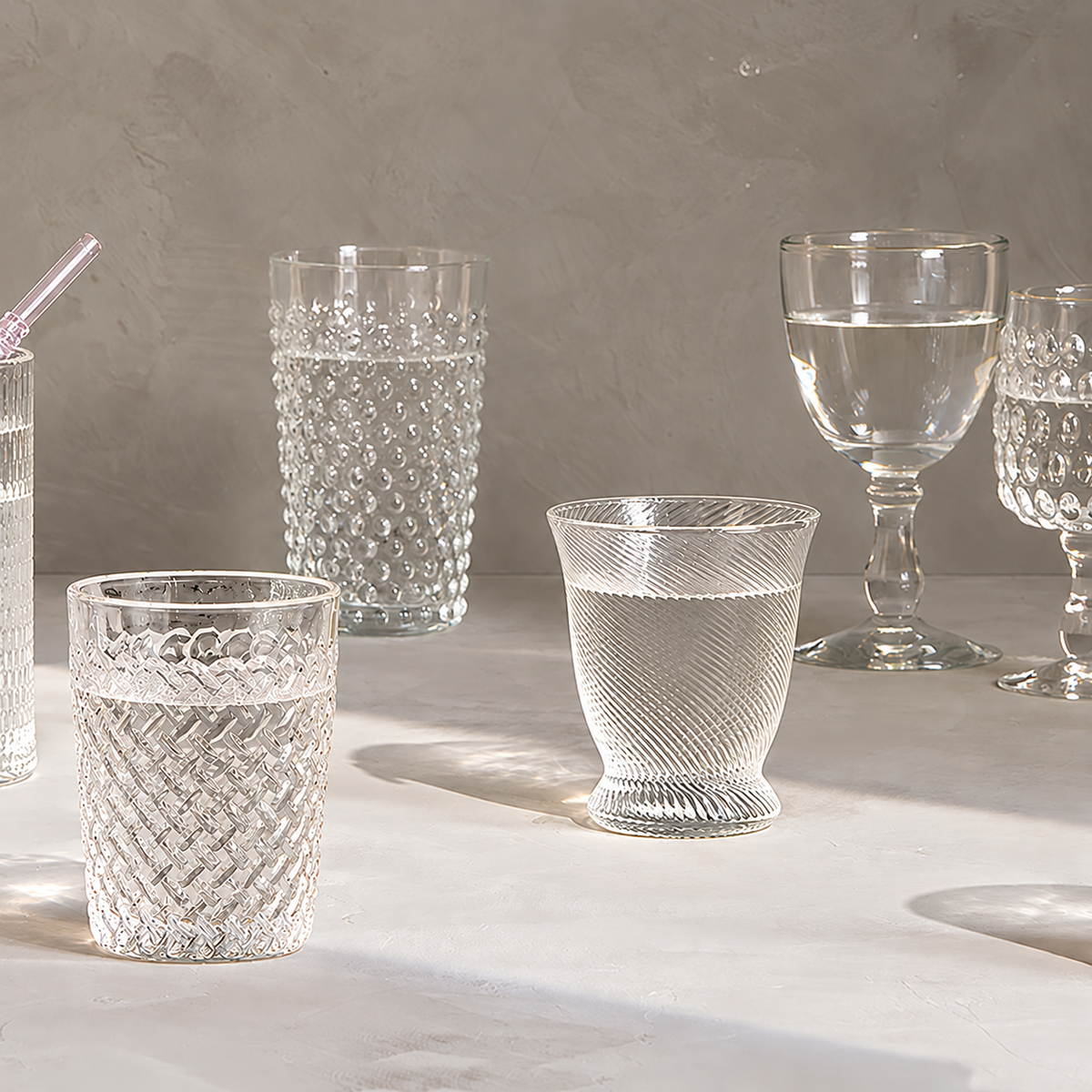 A collection of clear textured glass drinking vessels, including tumblers, a goblet, and a partially visible tumbler with a straw, arranged on a light stone surface against a neutral gray background.