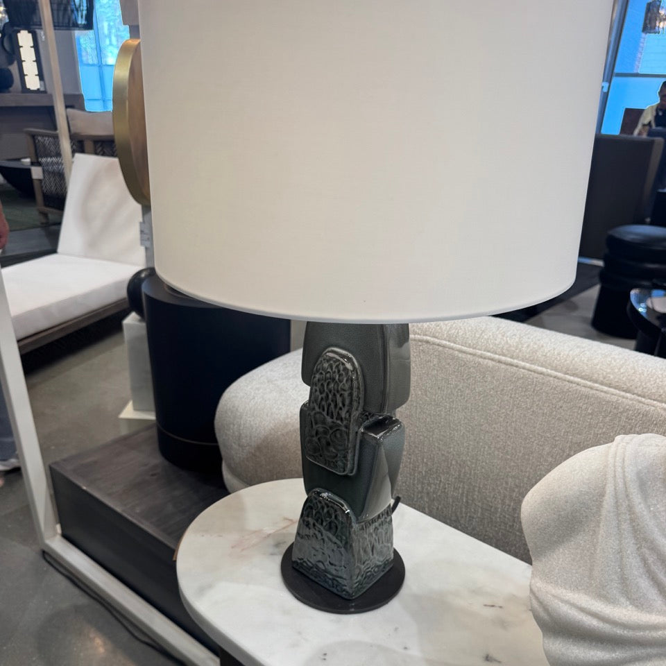 A modern table lamp with a large white drum shade and a dark textured ceramic sculptural base, displayed on a marble-topped side table in a furniture showroom.