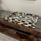A rectangular decorative tray with raised edges, featuring a mosaic pattern of black, white, tan, and brown stone-like pieces, displayed on a glass-topped wooden console table in an indoor setting.
