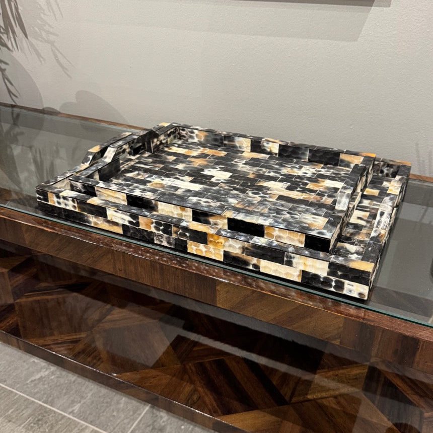 A rectangular decorative tray with raised edges, featuring a mosaic pattern of black, white, tan, and brown stone-like pieces, displayed on a glass-topped wooden console table in an indoor setting.