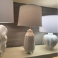 A table lamp with a textured white ceramic base and a drum shade, displayed on a shelf in a showroom alongside other lamps.