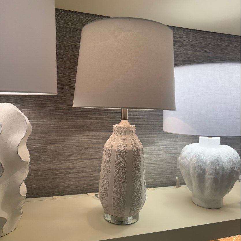 A table lamp with a textured white ceramic base and a drum shade, displayed on a shelf in a showroom alongside other lamps.