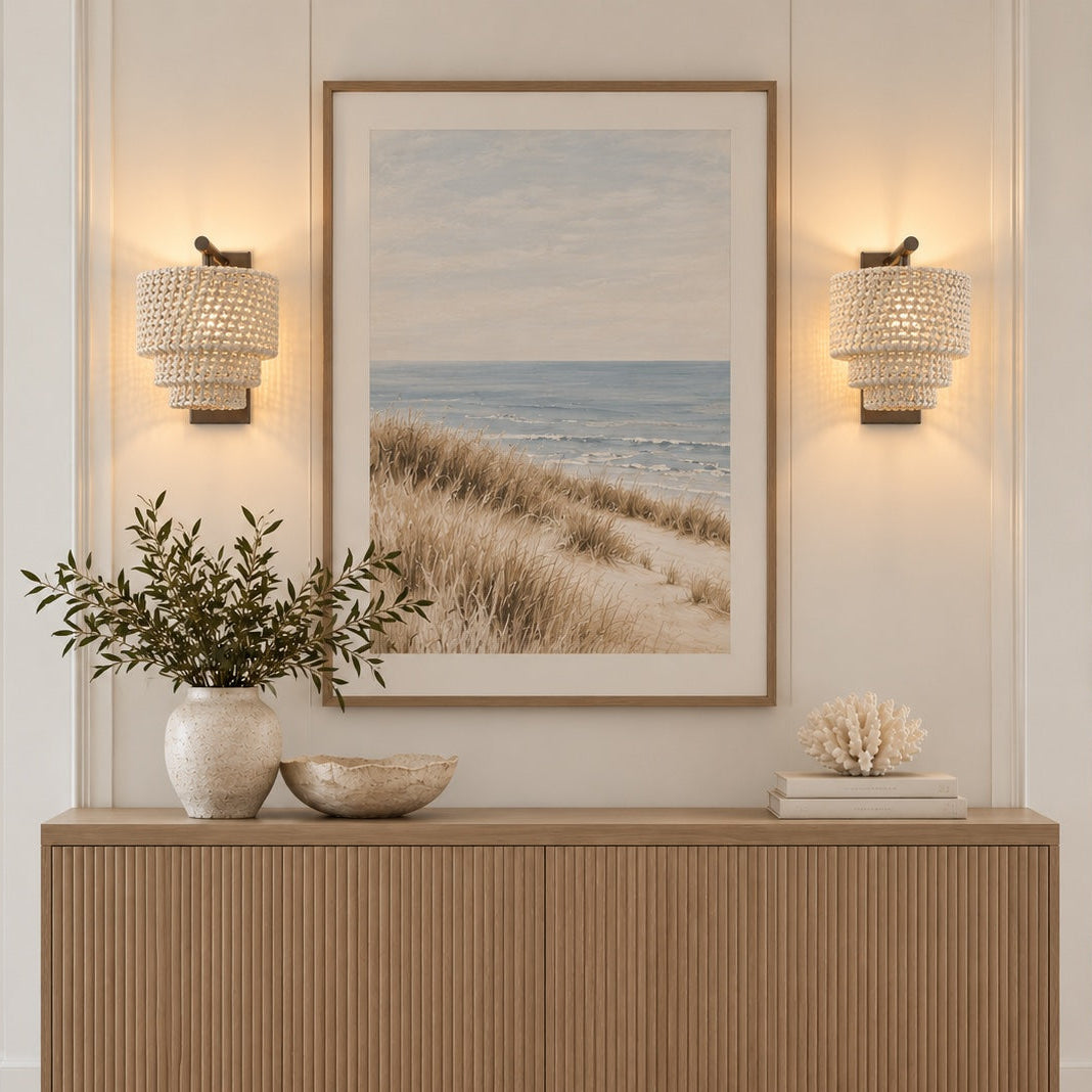 A coastal beach scene framed art print hanging on a white wall above a light wood console table, flanked by two lit woven wall sconces and styled with a vase of greenery and decorative bowls.