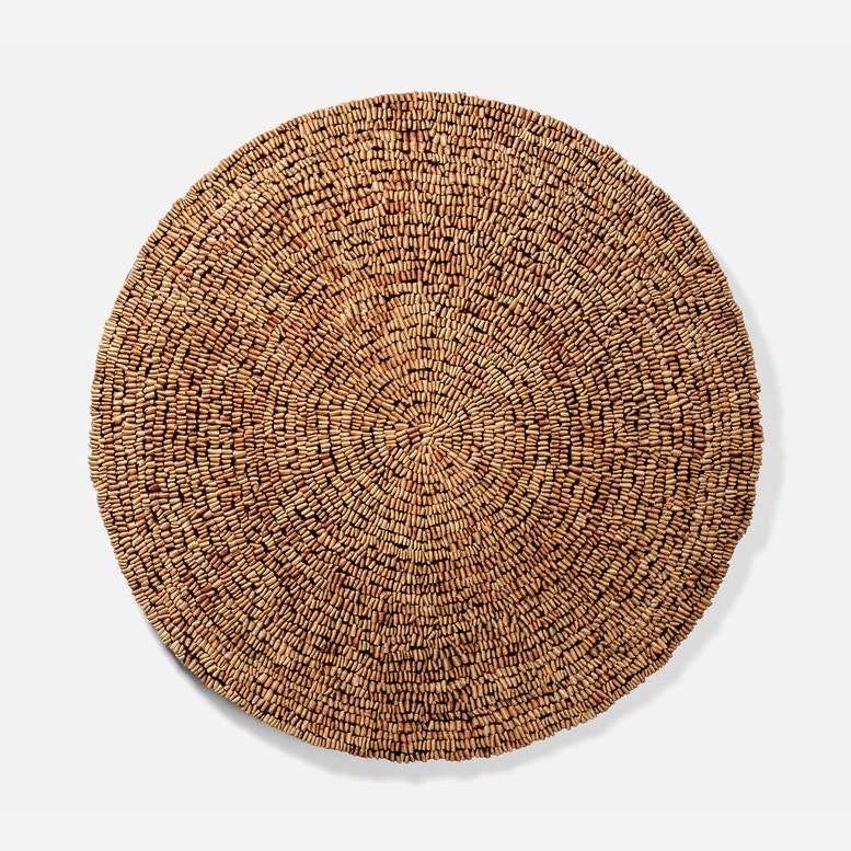 A round woven natural fiber placemat or rug in a warm tan-brown color, photographed from directly above on a light gray background.