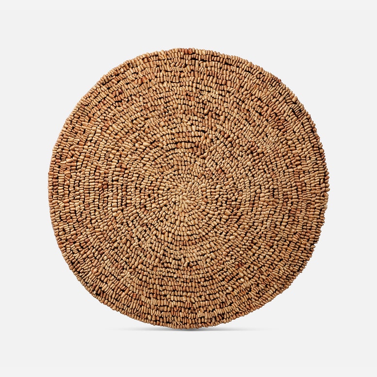 A round woven natural fiber placemat or rug in a light brown tan color, shown from a top-down view on a light gray background.