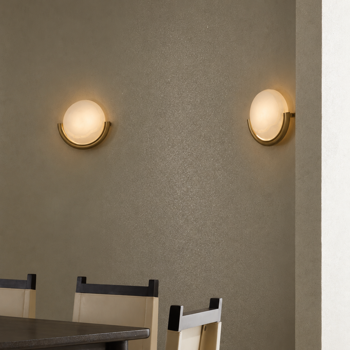 Two modern wall sconces with frosted oval glass shades and curved gold metal bases mounted on a textured beige wall above a dining table and chairs.