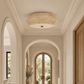 A round woven natural fiber ceiling light fixture installed in a bright, elegant hallway with arched doorways and warm wood trim.