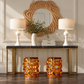 A styled interior console scene with two white table lamps, a woven round mirror, a vase with greenery, a small box, and two amber glass stool-like side tables on a dark marble-top console table against a beige paneled wall.