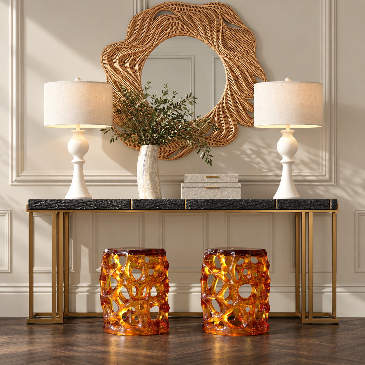 A styled interior console scene with two white table lamps, a woven round mirror, a vase with greenery, a small box, and two amber glass stool-like side tables on a dark marble-top console table against a beige paneled wall.
