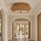 A round woven wicker ceiling light fixture with a natural tan finish, mounted on a white ceiling in a bright, modern hallway with arched doorways and light wood flooring.