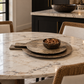 Elegant modern dining space featuring layered rustic stone serving boards and a matching bowl filled with mixed nuts on a white marble dining table, surrounded by woven rattan chairs with ivory cushions, black cabinetry, brass accents, and soft natural lighting.