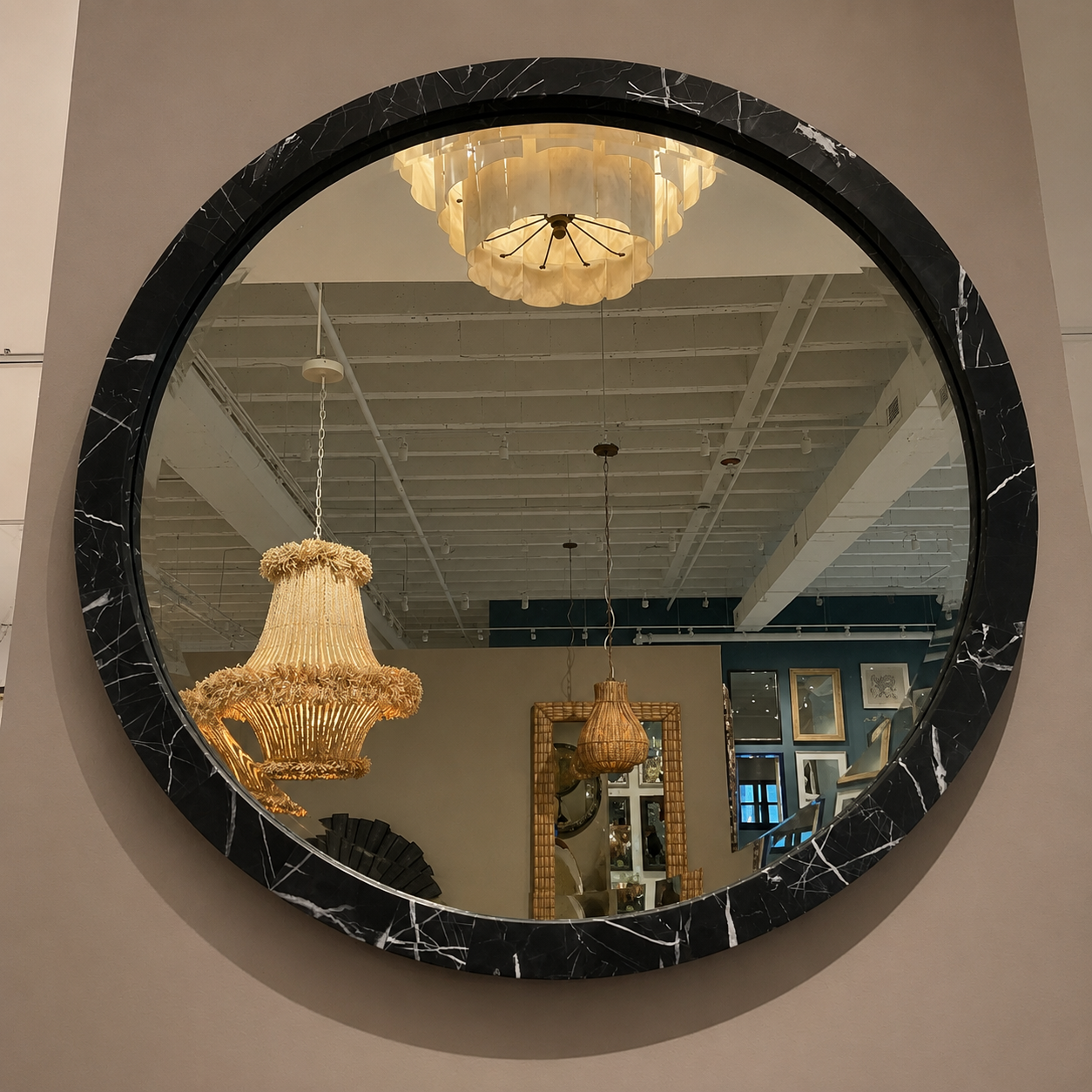 A large round wall mirror with a black marble-patterned frame, mounted on a beige wall. The mirror reflects a home decor showroom interior with hanging chandeliers and ceiling fixtures.