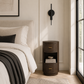A modern cylindrical nightstand in dark brown wood with gold-tone handles, styled next to a bed in a bright, contemporary bedroom.