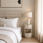A styled bedroom scene featuring a white two-drawer nightstand with gold handles next to a bed with white linens and a beige upholstered headboard. The nightstand holds a lit table lamp with a cream shade and gold base, set against a warm beige wall with a curtain on the right.