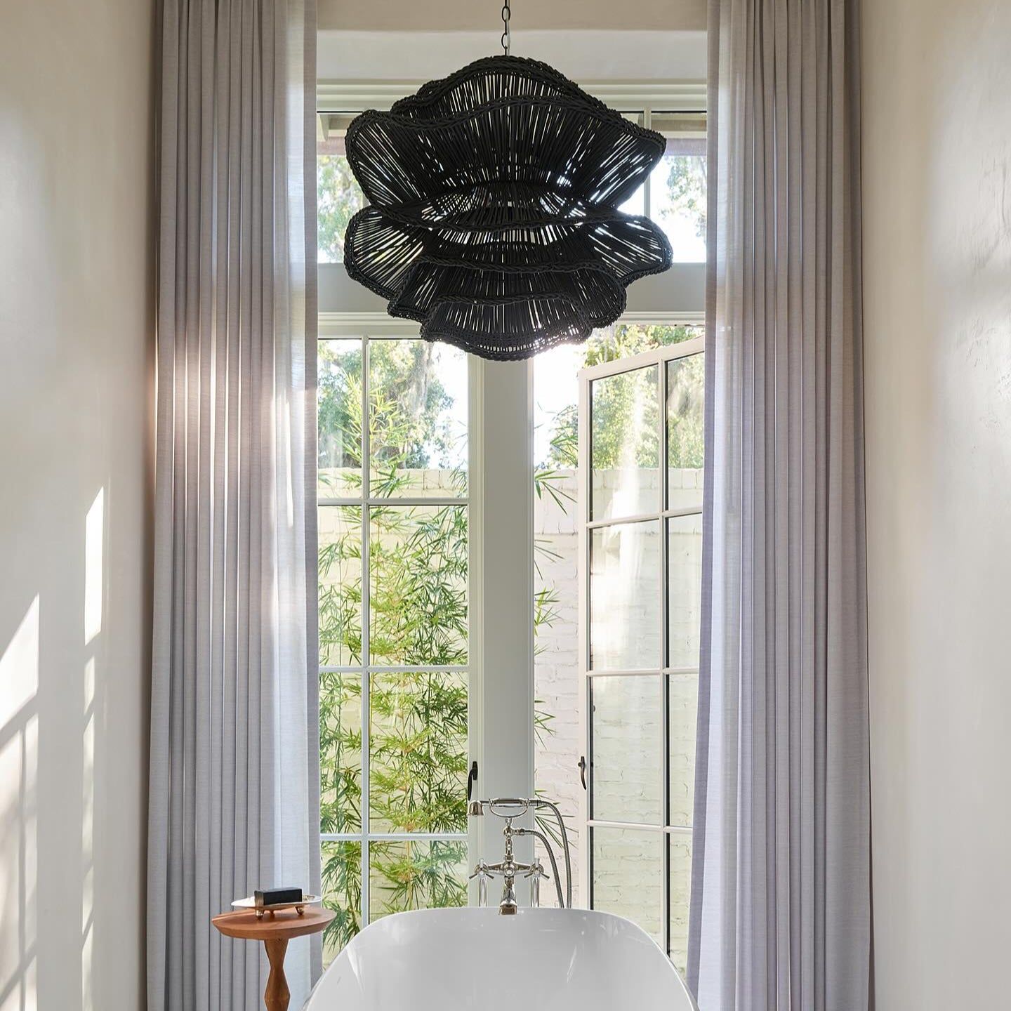 Bathroom with a freestanding white bathtub, black chandelier, and large windows with curtains.
